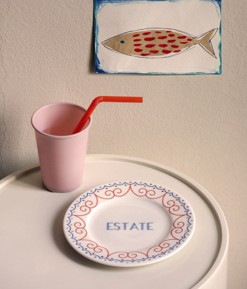 tavolino con bicchiere rosa con la cannuccia rossa e piatto in porcellana decorato con la scritta estate vicino a un muro beige con un disegno di un pesce 