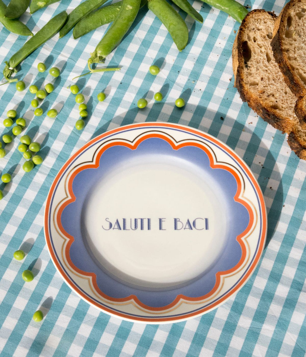 Piatto in porcellana con scritta saluti e baci su una tavola con tovaglia a quadretti azzurri e bianchi decorato con piselli e pane 