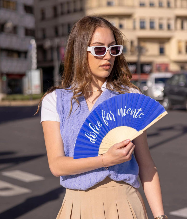 Giovane donna elegante in un contesto urbano tiene un ventaglio blu con la scritta "dolce far niente" in caratteri bianchi. Indossa occhiali da sole bianchi, un top senza maniche blu e una gonna plissettata beige.