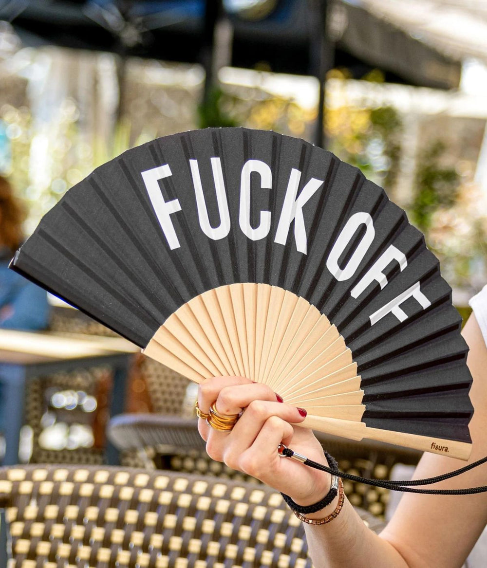 Una mano femminile tiene un ventaglio aperto con la scritta "FUCK OFF" in un caffè all'aperto, evidenziando un modo audace e spiritoso di godersi l'estate.