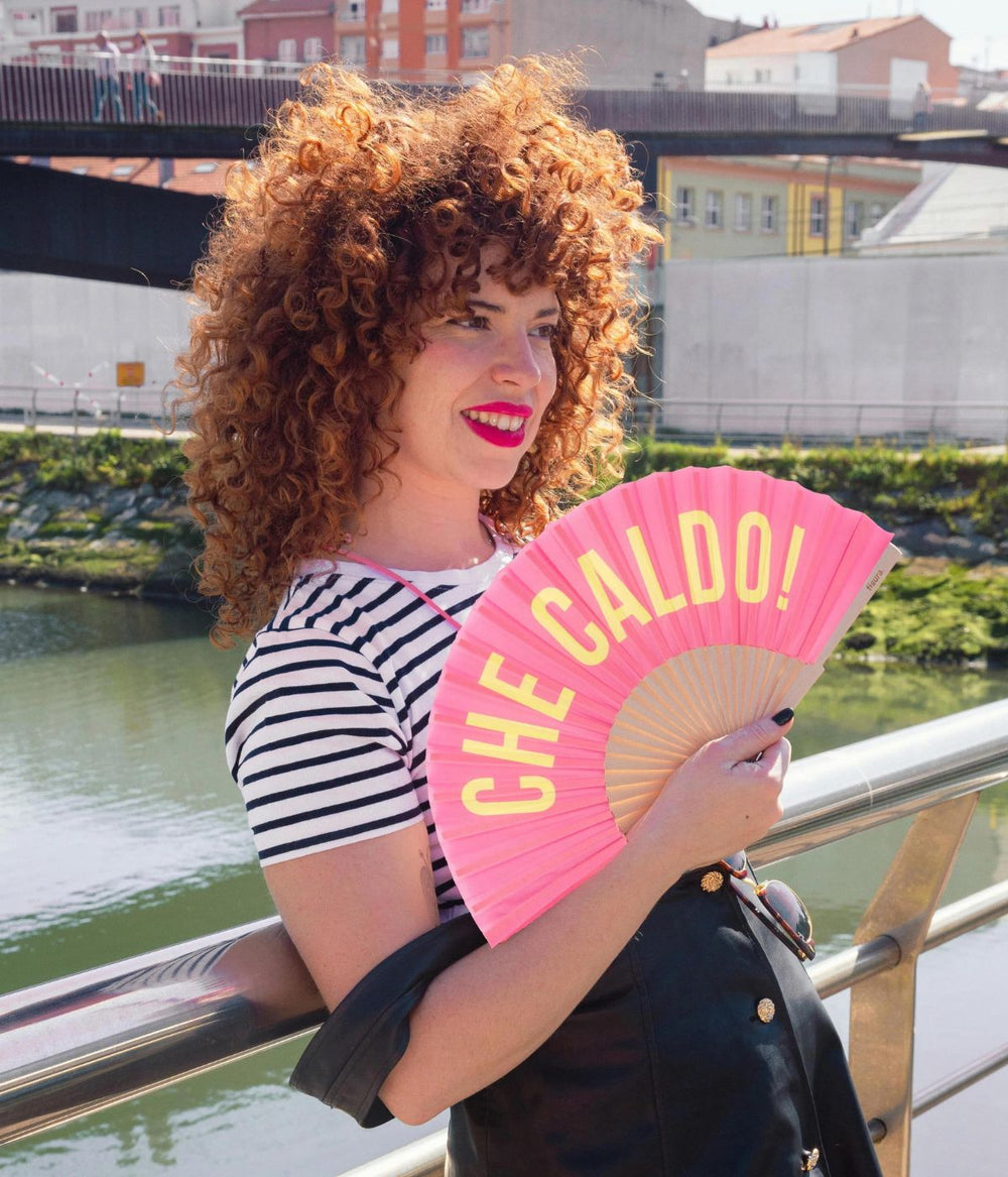 Giovane donna con ricci capelli rossi sorride mentre tiene un ventaglio rosa con la scritta "CHE CALDO!" su sfondo urbano con fiume.