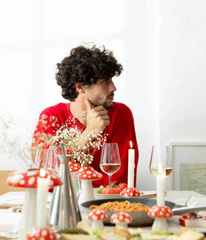 Un uomo con capelli ricci seduto a una tavola decorata con funghi ornamentali a pois rossi e bianchi, candele accese e piatti di cibo, incluso un piatto di spaghetti al pomodoro.