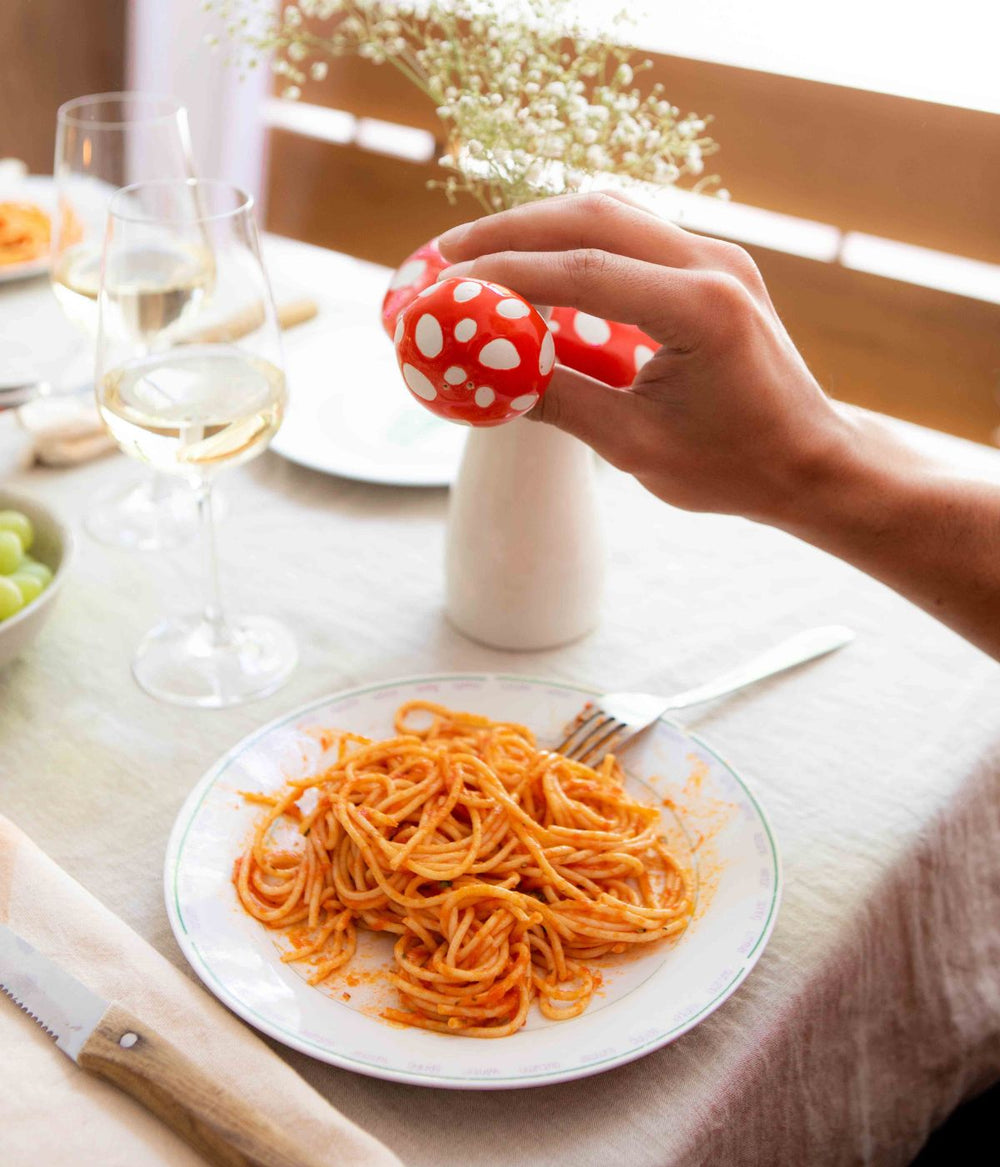 Una mano che tiene una saliera a forma di fungo a pois rossi e bianchi sopra un piatto di spaghetti al pomodoro su un tavolo apparecchiato con bicchieri di vino bianco e decorazioni floreali.
