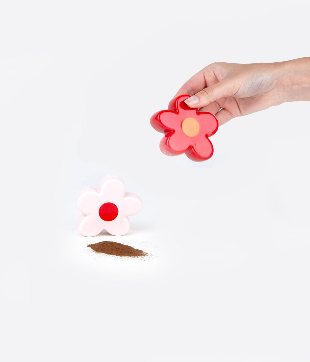 Una mano tiene uno spargipepe rosso a forma di fiore, facendo cadere pepe sul tavolo. Accanto c'è un altro spargipepe bianco a forma di fiore, entrambi su uno sfondo bianco.
