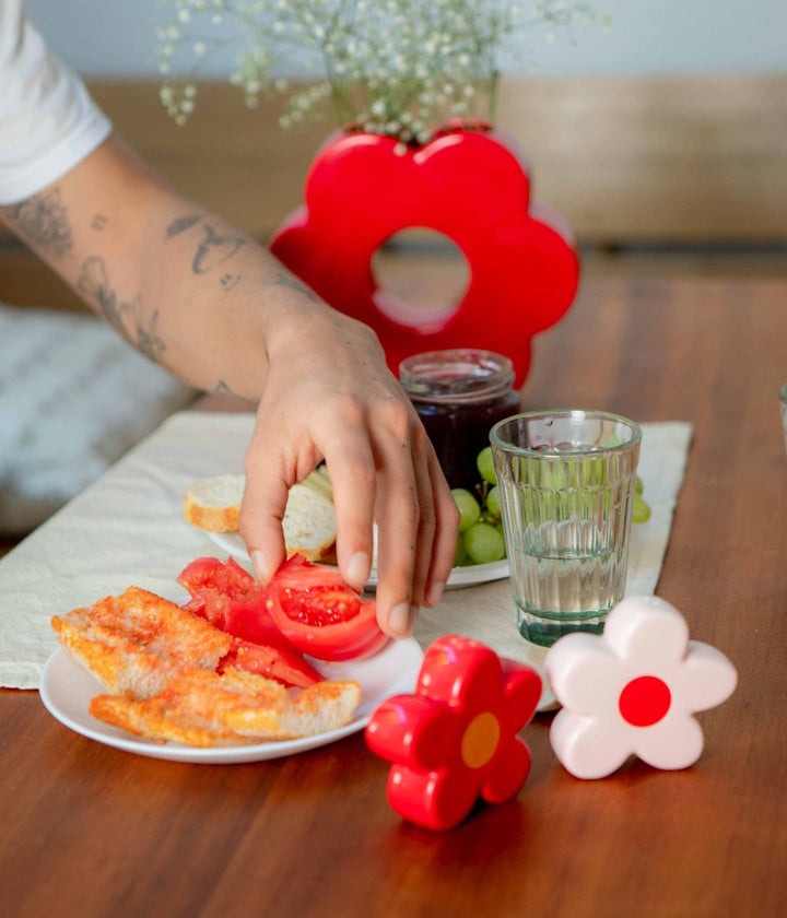 Una mano sta prendendo un pezzo di pomodoro da un piatto con uova fritte e pomodori a fette. Sul tavolo sono presenti bicchieri d'acqua, un vaso con fiori bianchi e un portafiori rosso a forma di fiore, oltre a due spargipepe a forma di fiori colorati.