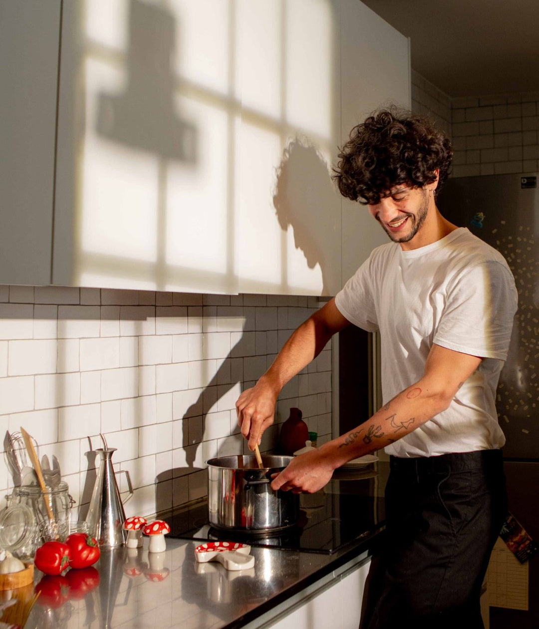 Un uomo con capelli ricci e una maglietta bianca mescola una pentola in cucina. Sul piano di lavoro ci sono utensili da cucina e decorazioni a forma di funghi rossi a pois bianchi.