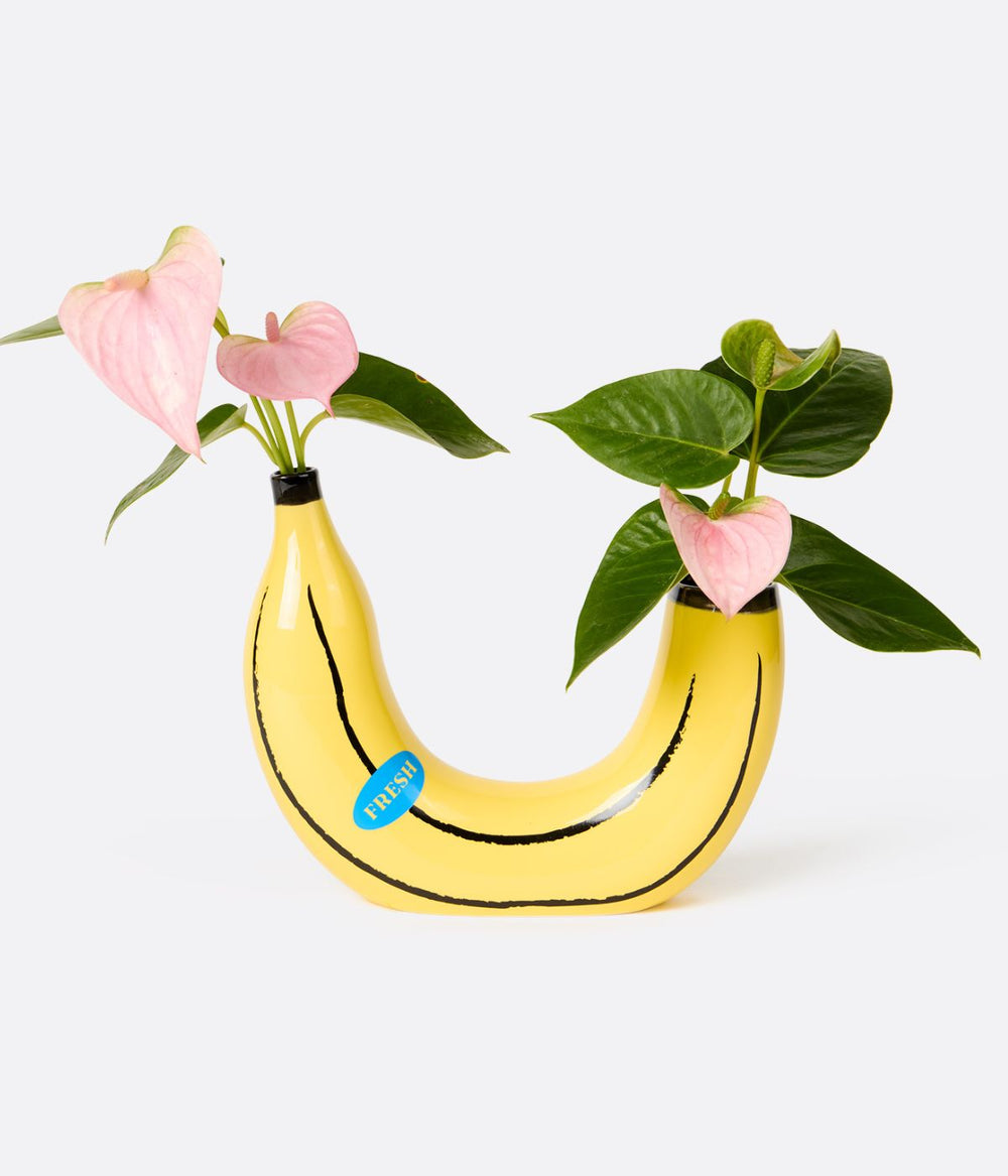 Vaso a forma di banana gialla con due spazi per fiori, contenente fiori rosa e foglie verdi, su sfondo bianco.