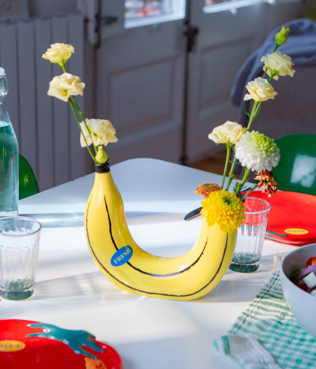 Vaso a forma di banana gialla con due spazi per fiori, contenente fiori gialli e bianchi. Il vaso è posizionato su un tavolo apparecchiato con bicchieri, piatti e altri utensili da cucina.