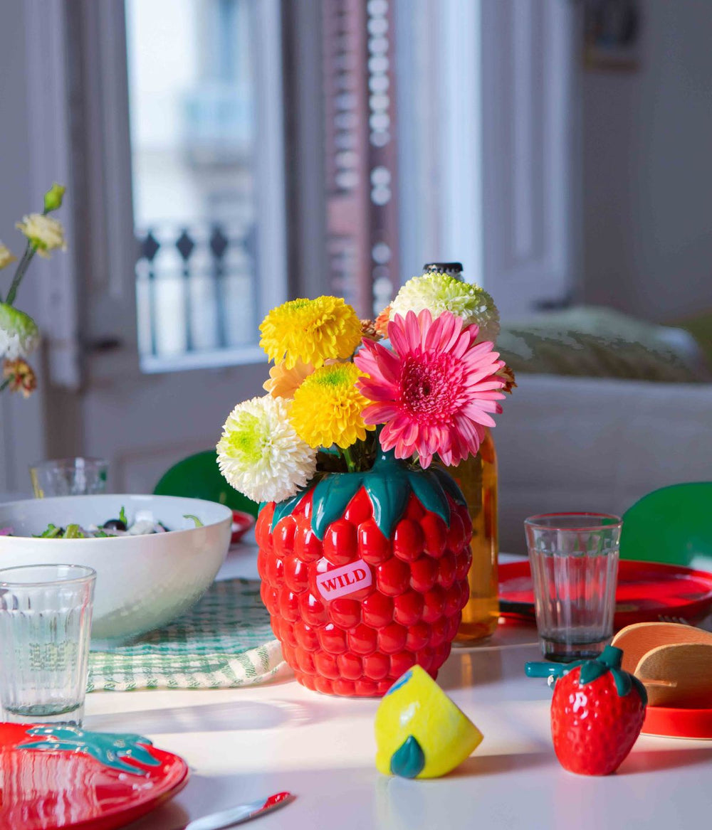 Un vaso a forma di lampone rosso con dettagli verdi, contenente vari fiori colorati, posizionato su un tavolo apparecchiato con bicchieri, piatti e altri utensili da cucina, in una stanza luminosa con porte-finestre sullo sfondo.
