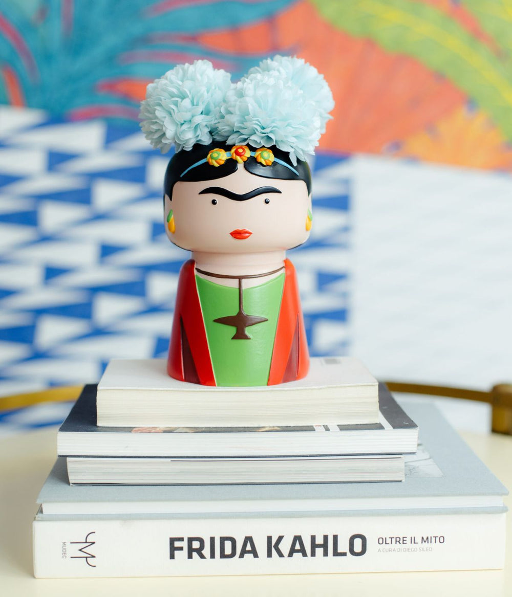 Un vaso in poliresina a forma di Frida Kahlo, con fiori azzurri tra i capelli, posizionato su una pila di libri, con uno sfondo colorato e geometrico.