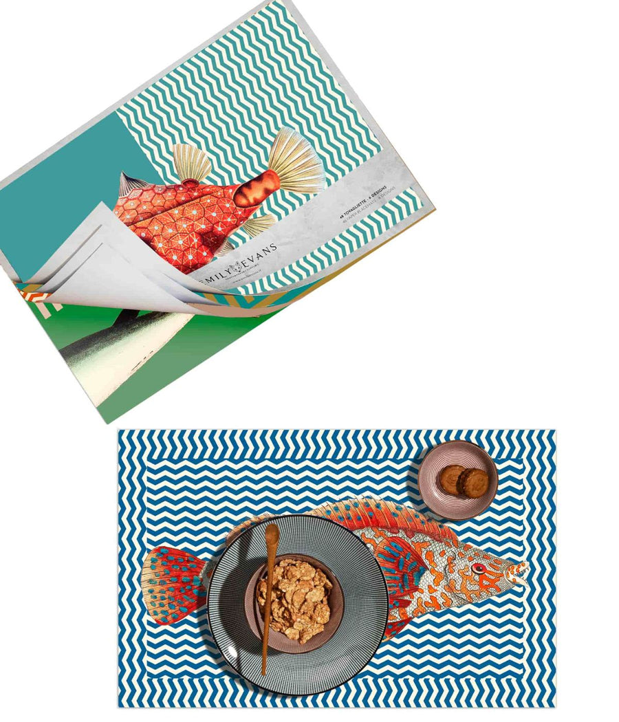 Composizione creativa con tovagliette americane a tema marino con motivi a zigzag e illustrazioni di pesci colorati, accompagnata da una colazione su piatti moderni
