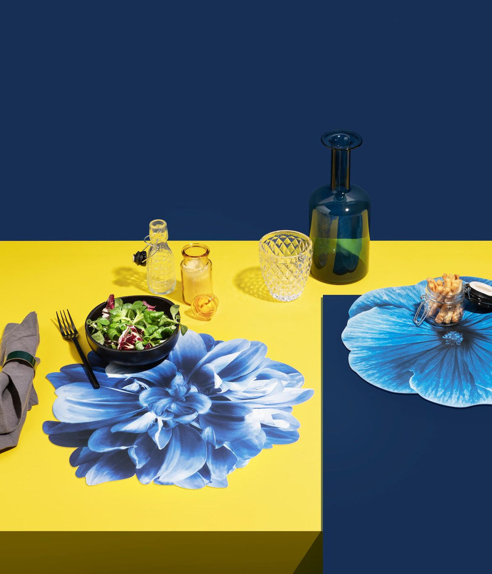 Tavola elegante con tovaglietta americana blu con disegno di un grande fiore, accompagnata da stoviglie e decorazioni floreali in un'ambientazione vivace.
