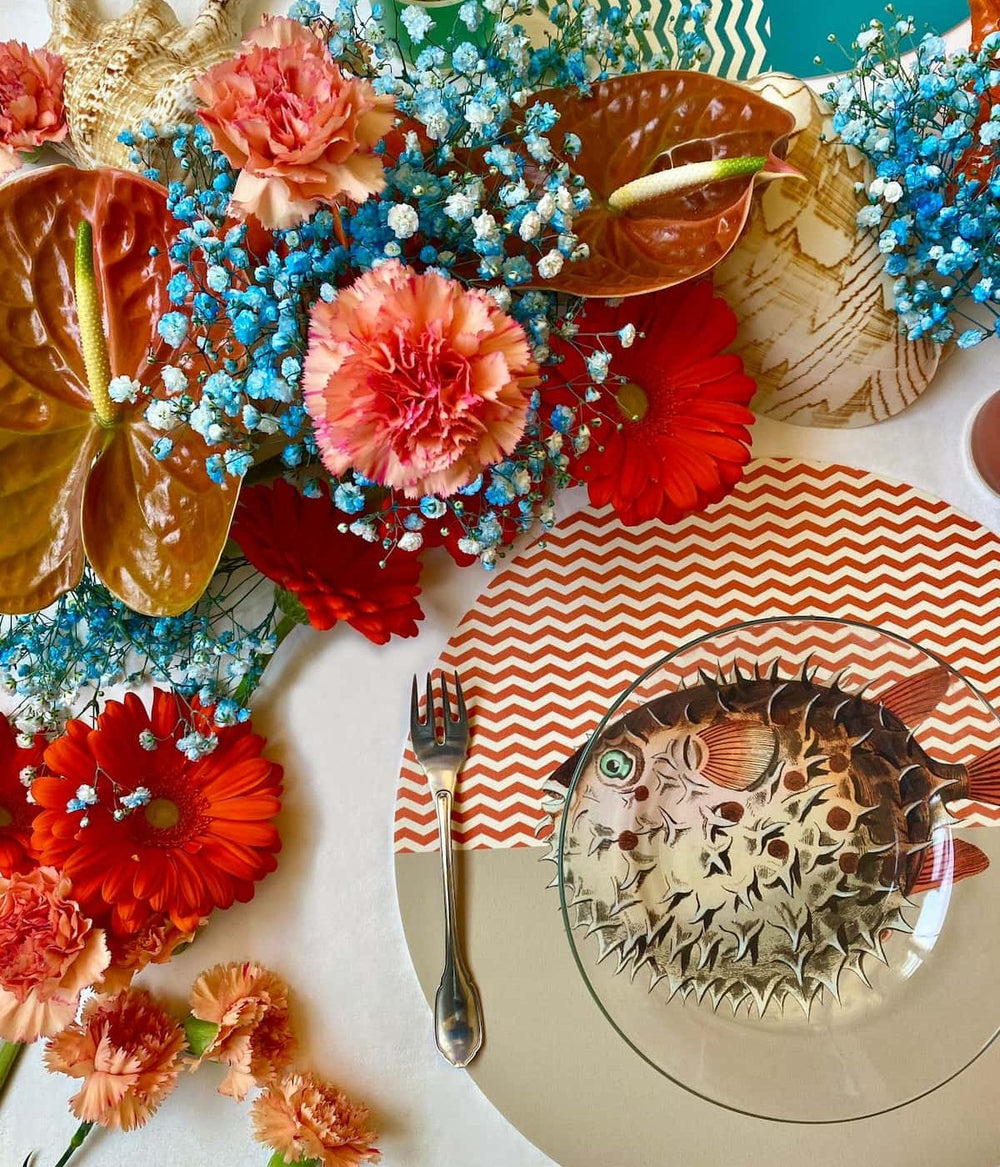 avola da pranzo estiva decorata con un piatto trasparente che mostra una tovaglietta americana rotonda con llustrazione di un pesce palla, circondato da fiori vivaci e piatti decorativi.