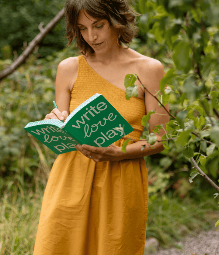 Donna con abito giallo che scrive sul Notebook verde con scritta Write love play in un giardino.