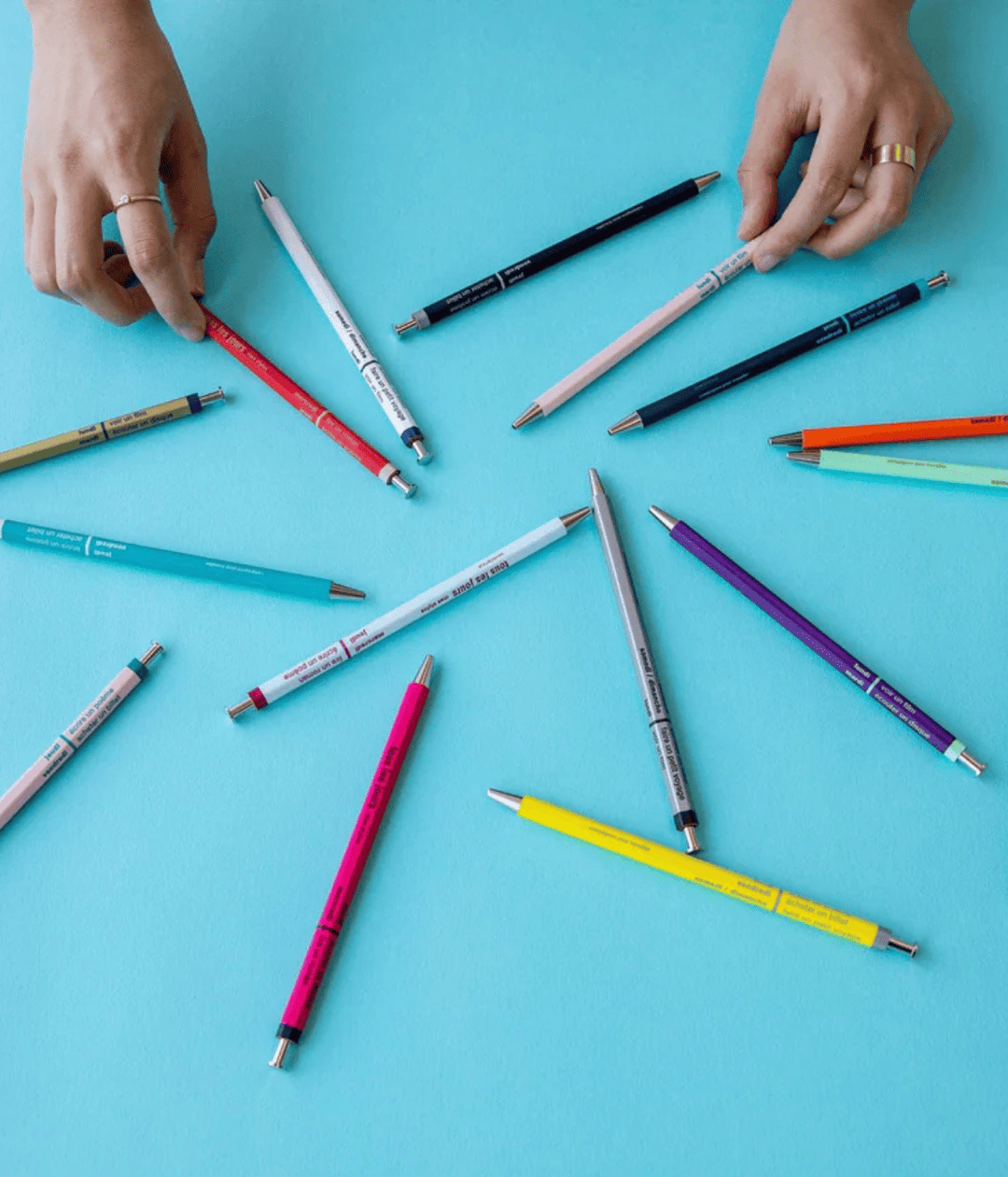 Penne di diversi colori appoggiate su tavolo azzurro. Due mani che tengono due penne. 